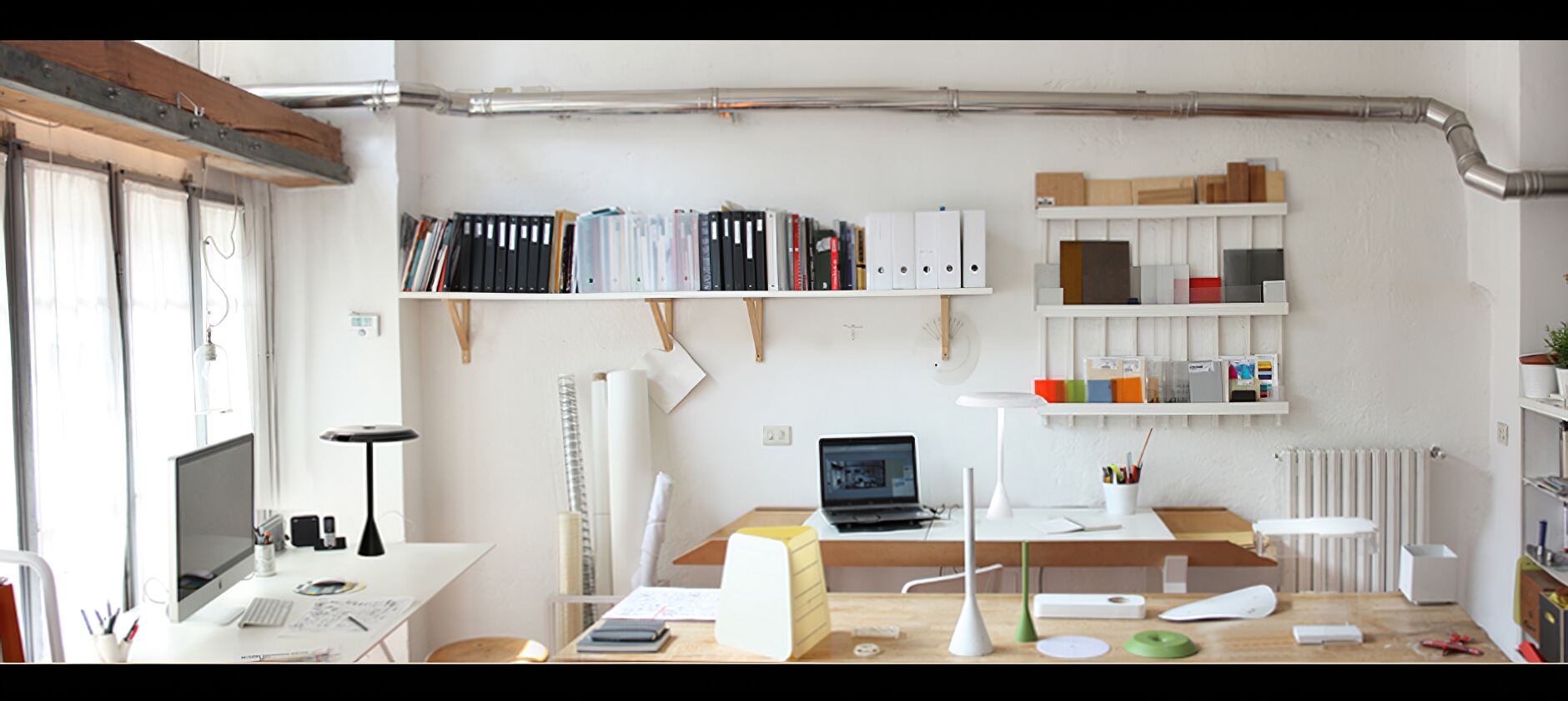 Heller Arbeitsbereich eines Designstudios mit grossen Fenstern, Schreibtischen, Regalen voller Ordner und Materialmuster sowie verschiedenen Designobjekten und Leuchten auf einem zentralen Arbeitstisch.
